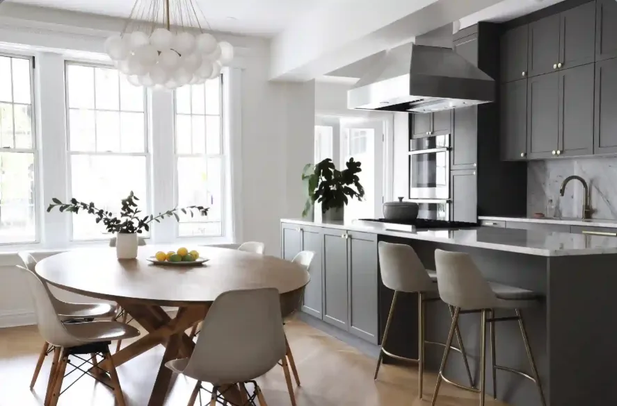 A modern kitchen with a wooden dining table and gray cabinets. Bright windows illuminate the space.